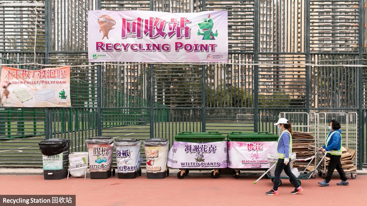 Recycling station at a Green Lunar New Year fair with recycling collection bins for waste paper, metals, plastic bottles, wilted plants, peach blossom trees and polyfoam boxes, and a general waste bin, also there are two cleansing workers on duty