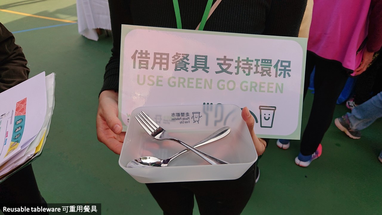 一名工作人員手持一個指示牌和一個食物托盤上面放了不銹鋼叉及匙羹，她正在宣傳可重用餐具借用服務